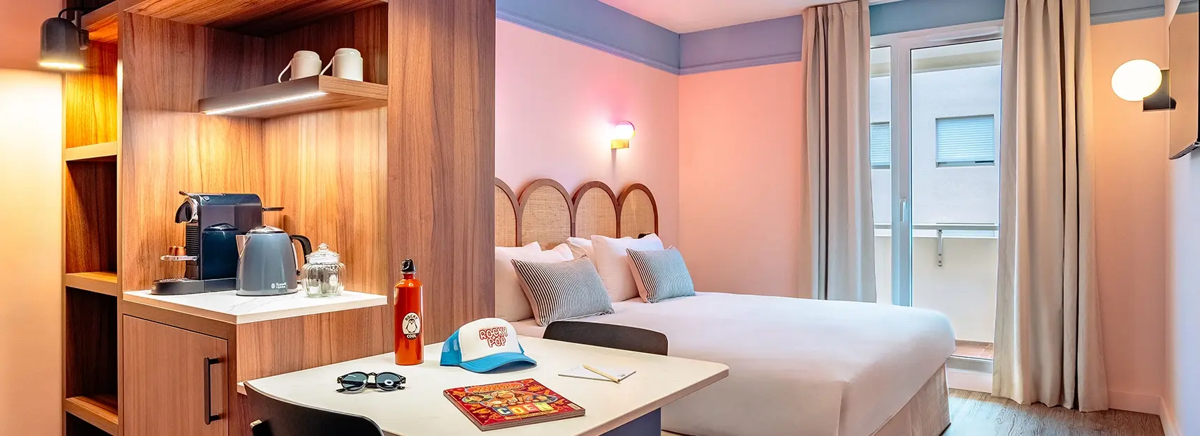 Chambre avec lit double au centre, petite table avec deux chaises à gauche, coin cafetière et bouilloire, grande fenêtre en fond – RockyPop Marseille
