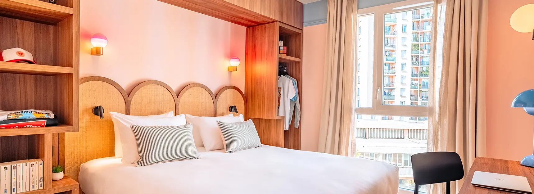Vue sur une chambre avec lit à gauche et rangement autour du lit, bureau à droite et une grande fenêtre au fond - rockypop marseille