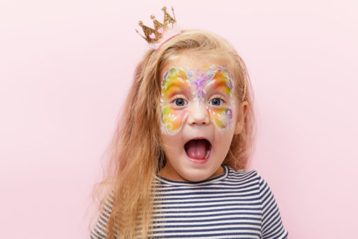 Pretty exciting smiling blond little child girl of 3 years with a bright face painting on pink background. Fun emotions, shocked face